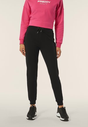 Pantalons de survêtement noirs avec des poignets élastiques et une taille à cordon de serrage, associés à un sweat-shirt court rose vif avec un texte de logo blanc. Des baskets noires complètent le look.
