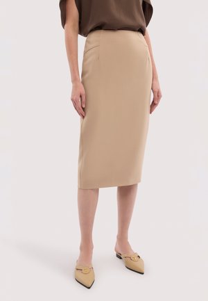 Kvinde iført beige knælængde blyantskjørt, brun løs top og beige mulepumps med spidse tæer og gulddetaljer, stående på hvid baggrund.
