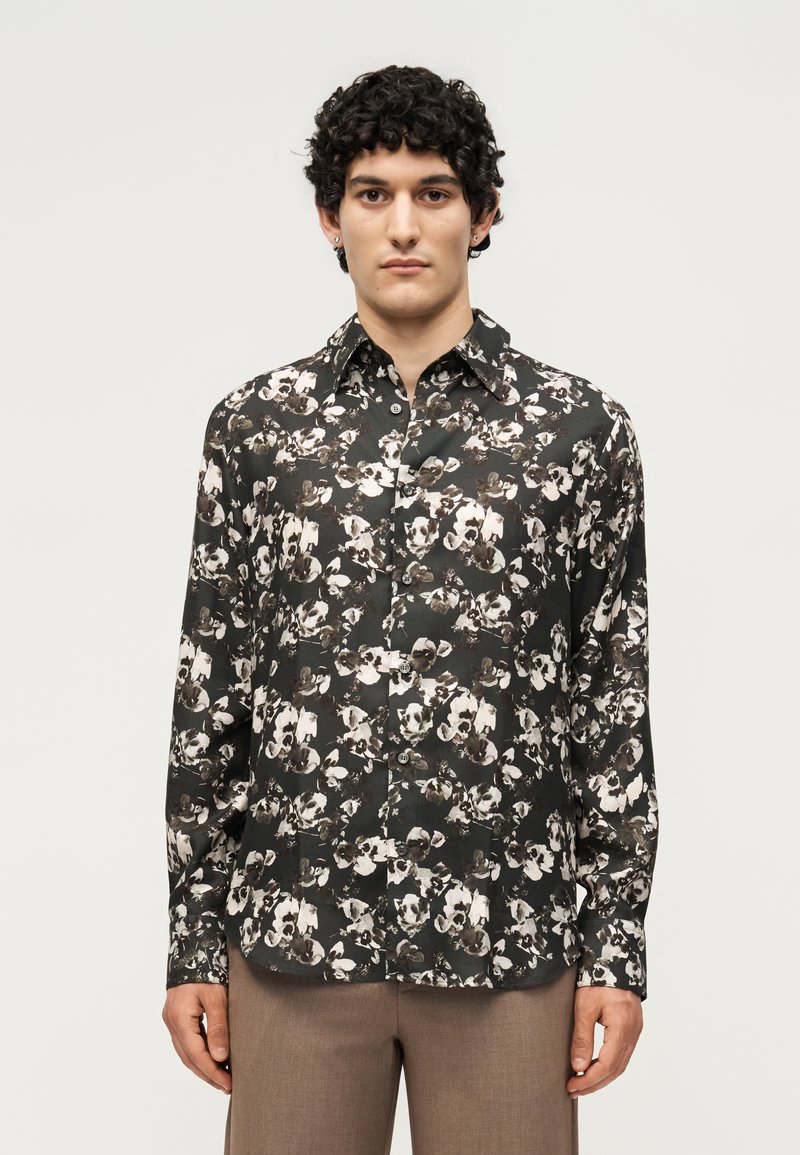 Νέος άνδρας που φορά μαύρο πουκάμισο με κουμπιά μακριάς μανίκιας με λευκά και μπεζ ανθισμένα σχέδια, συνδυασμένο με καφέ παντελόνι, στέκεται μπροστά σε ένα απλό φόντο.