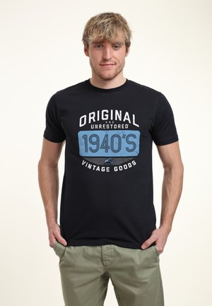 Jeune homme aux cheveux blonds portant un t-shirt noir avec l'inscription "Original et Non Restauré - Produits Vintage des années 1940" et un pantalon vert olive.