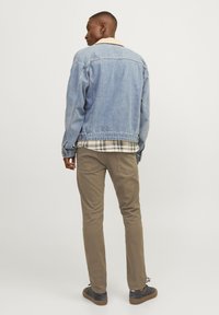 Uomo in piedi di spalle, che indossa una giacca di denim chiaro, una camicia a scacchi, pantaloni marroni e sneakers scure su uno sfondo uniforme.