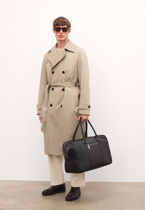 Bež trench coat mustrite musta nuppe, vöö ja küljetaskud, paigutatud musta nahkrahakoti ja tumepruunide saapadega. Prillid peas.