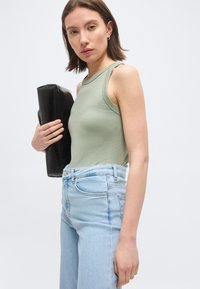 Νεαρή γυναίκα με κοντά καστανά μαλλιά, που φοράει αμάνικο ανοιχτό πράσινο τοπ και ανοιχτό μπλε τζιν, κρατώντας μια μαύρη τσάντα φάκελο κάτω από το χέρι της.