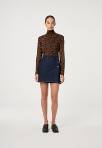Braun gepunktetes Turtleneck-Oberteil mit hohem Kragen, kombiniert mit einem dunklen Denim-Minikleid, das über einen seitlichen Knopfverschluss verfügt. Schwarze Slipper.