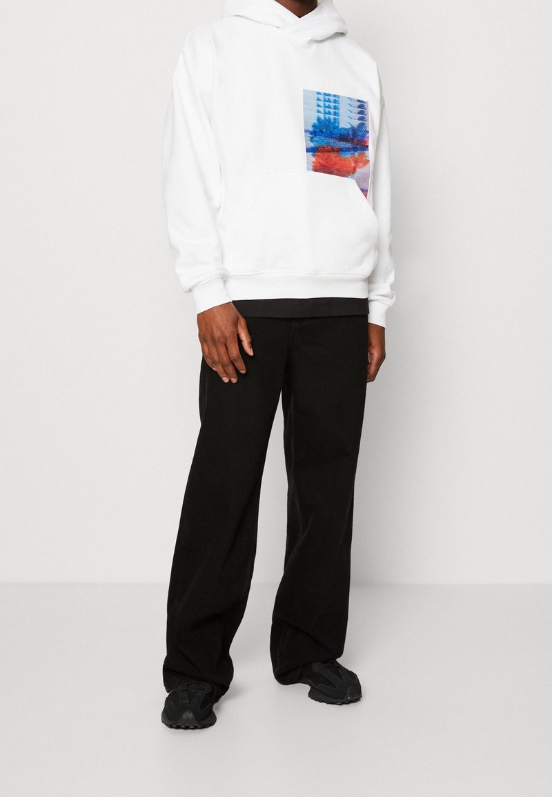 Felpa bianca con tasca frontale, caratterizzata da un design grafico colorato. Abbinata a pantaloni neri a gamba larga e sneaker nere.