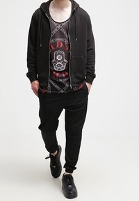 Černá mikina na zip přes vzorované tílko s červenými akcenty. Černé kalhoty a tenisky dotvářejí celý outfit. Zaměření na minimalistický design.