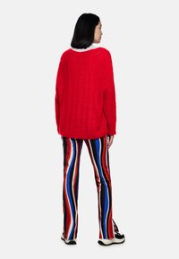 Roter Strickpullover mit lässiger Passform, kombiniert mit bunten gestreiften Schlaghosen in Rot, Blau, Weiß und Schwarz. Sneaker mit dicken Sohlen.