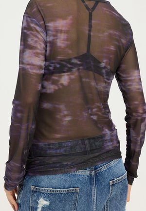 Durchscheinendes, langärmliges lila Top mit Muster, das einen schwarzen BH darunter zeigt, kombiniert mit ausgewaschenen blauen Jeans.