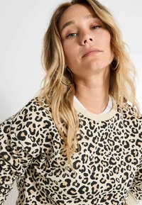 Sweat-shirt beige avec un imprimé léopard noir. Fabriqué en tissu doux avec un col en côtes et des manches longues.