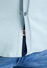 Camicia azzurra chiara con texture e dettagli in bottone, caratterizzata da un accento blu navy all'orlo e un'etichetta visibile all'interno della camicia.