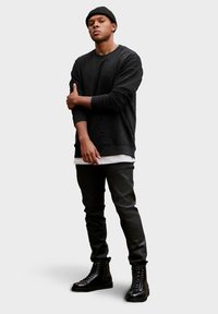 Felpa nera con texture e colletto tondo, indossata sopra una camicia bianca, abbinata a pantaloni neri slim-fit e stivali neri con lacci.