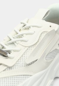 Hvit og grå mesh sneakers med hvite lisser, polstret krage, teksturert såle, og en metallisk snørelapp på venstre frontside.