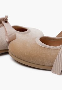 Ballerines beige en tissu doux avec un texture veloutée. Elles présentent une ouverture ronde et une sangle en ruban assortie sur le côté.