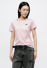 Rózsaszín v-nyakú póló puha anyagból, bal mellrészen egy kis kék "HUGO" logóval, sötétszürke, kopottas farmerrel és fekete övvel párosítva.