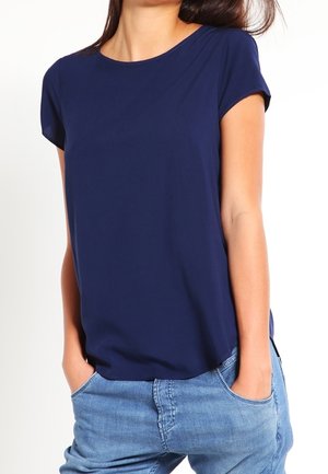 Vrouw draagt een effen donkerblauw T-shirt met korte mouwen en lichtblauwe spijkerbroek met handen in de zakken, tegen een witte achtergrond.