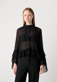 Kvinna i en genomskinlig svart blus med volanger och utsvängda ärmar över en svart bandeau, kombinerat med svarta byxor och stora hoopörhängen.