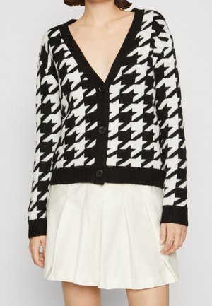 Cardigan pied-de-poule noir et blanc avec boutons, associé à une jupe plissée blanche porté par une personne sur un fond uni.