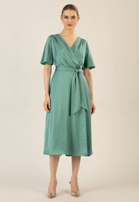 Un vestito avvolgente in raso teal con maniche a sbuffo corte, scollo a V e vita con lacci. La gonna arriva a metà polpaccio, con una texture liscia.