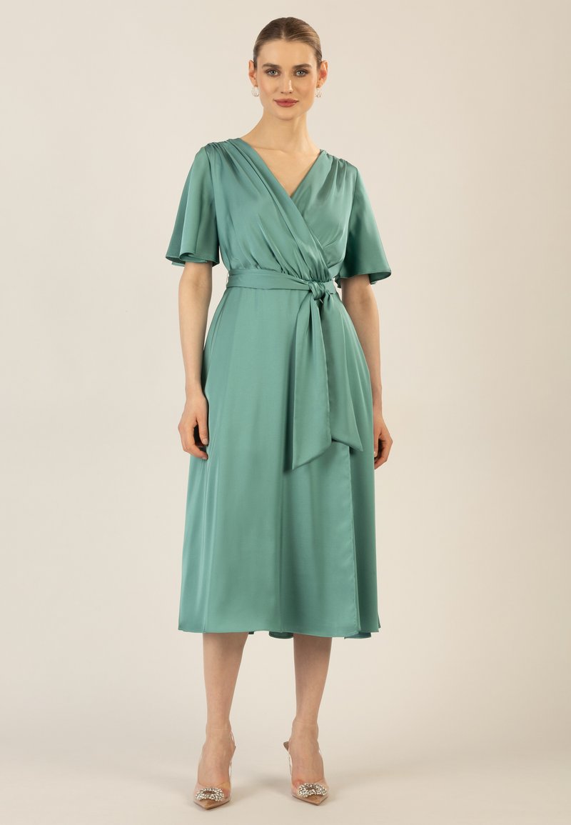 Un vestito avvolgente in raso teal con maniche a sbuffo corte, scollo a V e vita con lacci. La gonna arriva a metà polpaccio, con una texture liscia.