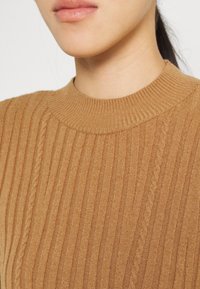 Gestrickter Rippenpullover in Hellbraun mit hohem Kragen und strukturiertem Muster. Das Material wirkt weich mit subtilen Farbvariationen.