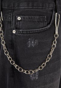 Črne denim hlače z raztrgano žepom in kovinsko verigo pritrjeno na zanko za pas, s srebrnim okovjem in krožnimi povezavami.