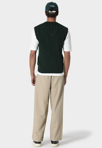 Vihreä palmikkoneuleliivi, hihaton malli, paksu rakenne, yhdistetty valkoiseen lyhythihaiseen paitaan ja beigeihin housuihin. Rentoon käyttöön tarkoitetut kengät.