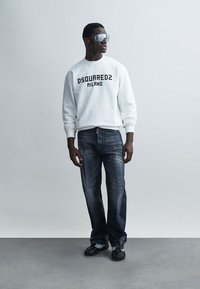 Sweat-shirt blanc avec "DSQUARED2 MILANO" en police noire, associé à un jean bleu foncé et des chaussures noires, accompagné de lunettes de soleil sportives.