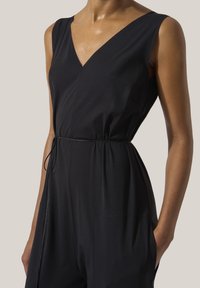 Ärmlös svart jumpsuit med V-ringning och smalt bälte i midjan, bärs av en person med en hand i fickan framför en enfärgad bakgrund.