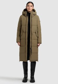 Cappotto lungo imbottito verde oliva con cappuccio, dotato di chiusura con zip e bottoni a scatto, e tasche laterali, indossato sopra un outfit nero e stivali al ginocchio.