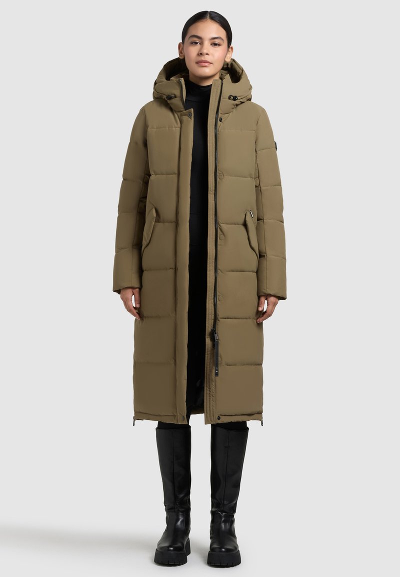 Cappotto lungo imbottito verde oliva con cappuccio, dotato di chiusura con zip e bottoni a scatto, e tasche laterali, indossato sopra un outfit nero e stivali al ginocchio.