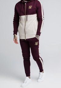 Ensemble de survêtement bordeaux et beige avec capuche zippée et logo. Présente un design bicolore, des poignets côtelés et des baskets blanches. Style simple et ajusté.