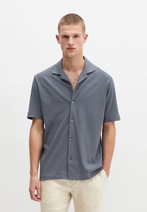 Młody mężczyzna z krótkimi włosami, ubrany w szarą koszulę z krótkim rękawem zapinaną na guziki i jasne spodnie, stojący na jednolitym białym tle.