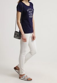 Camiseta navy con texto impreso, jeans skinny blancos, sandalias grises con hebillas y un bolso negro texturizado con cadena.
