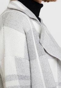 Frau in einer grau-weißen karierten Strickjacke über einem schwarzen Rollkragenoberteil, das an Hals und Schulter kurz geschnitten ist.
