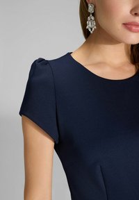 Marineblaues Kleid mit kurzen Ärmeln, aus glattem Stoff mit dezenten Falten. Auffällige Statement-Ohrringe mit klaren und silbernen Details.