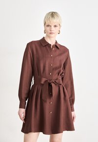 Hnedé ľanové šaty s dlhým rukávom, so zapínaním na gombíky vpredu, so zlatými akcentmi a viazaním v páse, s rozšírenou sukňou a golierom.