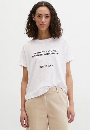Femme aux cheveux bouclés mi-longs portant un t-shirt blanc avec le texte "Respectez la nature, réécrivez demain depuis 1967" et un pantalon beige.