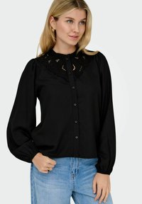 Giovane donna con capelli biondi, indossa una blusa nera con bottoni e dettagli in pizzo e jeans azzurri chiari, con una mano in tasca, che guarda a destra.