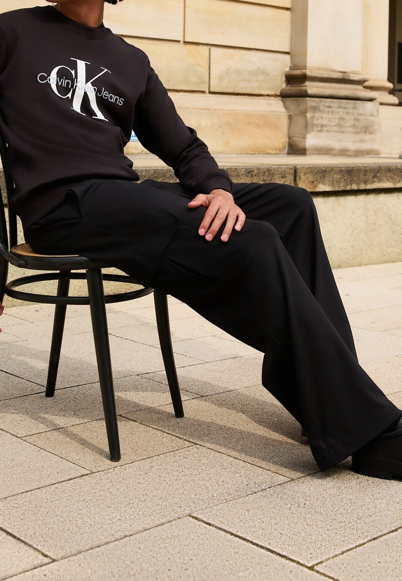 Personne assise sur une chaise noire en plein air, portant un sweat-shirt noir Calvin Klein Jeans et un pantalon noir ample avec des chaussures noires.