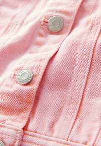 Heledad roosa denim kangas pehmea tekstuuriga, millel on hõbedased metallnupud ja nähtavad õmblused. Lähedalt vaade kangale ja riistvarale.