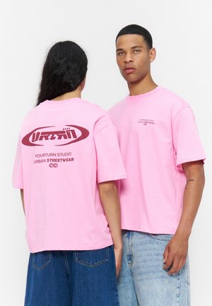 Deux personnes portent des t-shirts streetwear roses oversize ; l'une regarde devant, l'autre montre son dos avec un logo bordeaux et un texte.