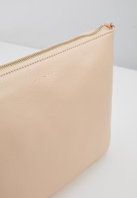 Pochette en cuir beige texturé avec une fermeture éclair dorée et le nom de la marque discrètement embossé sur une surface blanche.