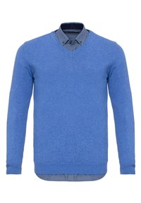 Pull bleu col en V superposé sur une chemise grise boutonnée dont le col est replié par-dessus le col du pull.