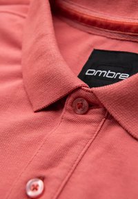Koralli polo-paita, jossa on teksturoitu kangas, terävä kaulus ja musta label, jossa lukee "Ombre". Sisältää napp closure, jossa on hienovarainen muotoilu.