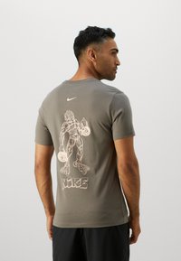 Rövid ujjú, szürke pamut póló, amelyen egy fehér grafikán izmos figura látható súlyokkal, és a hátoldalán "NIKE" felirat található.