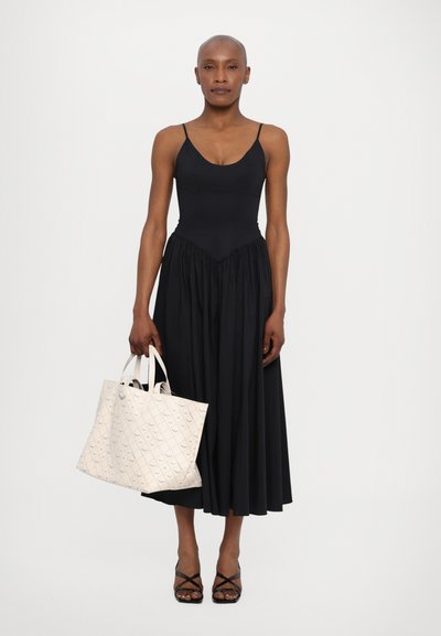 Robe noire sans manches avec un corsage ajusté et une jupe évasée, associée à un sac fourre-tout texturé crème et des sandales noires à lanières.