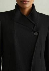 Cappotto in lana nera con colletto alto, caratterizzato da una chiusura a bottoni diagonale e linee pulite, che mette in mostra un tessuto a trama liscia.