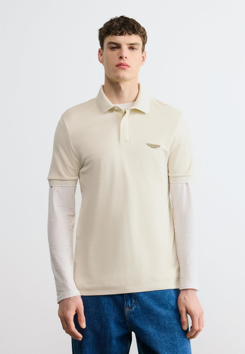 Νεαρός άνδρας που φοράει μπεζ κοντομάνικο πόλο πάνω από λευκό μακρυμάνικο πουκάμισο και μπλε τζιν, στέκεται μπροστά σε απλό φωτεινό φόντο.