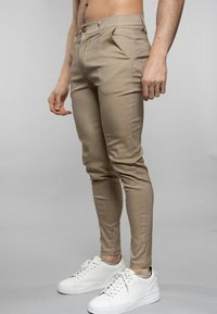 Pantalons beiges ajustés avec des plis, fabriqués à partir d'un tissu lisse. Associés à des baskets blanches de sport, présentant un design épuré.