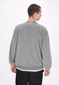 Šedá oversized mikina s dlouhými rukávy, žebrovanými manžetami a lemem. Měkká textura s ležérním střihem. Nositá přes bílou vrstvenou košili.